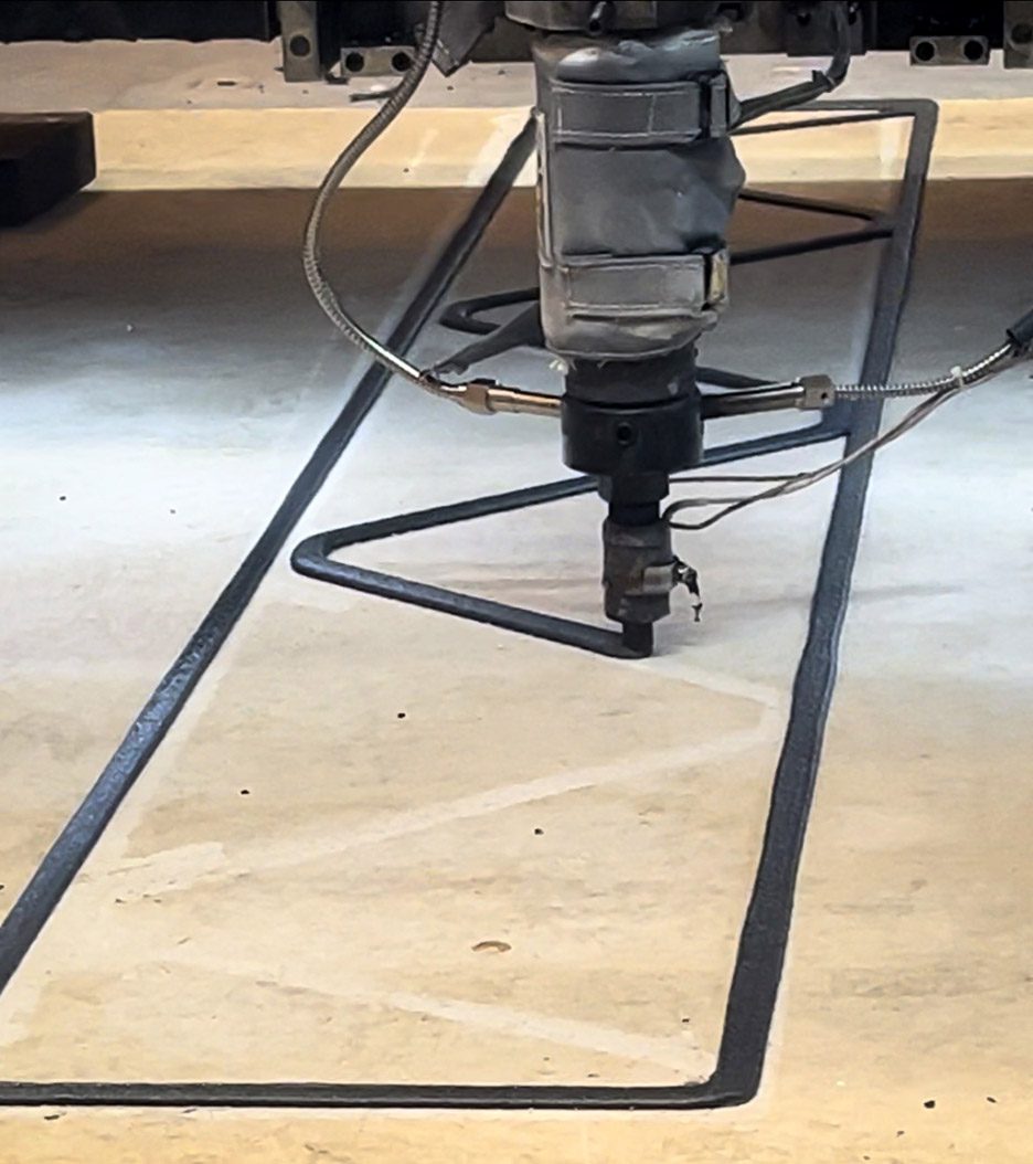 Photo of a large-scale 3D-printing nozzle extruding a dark plastic-glass composite material into a zigzag shape